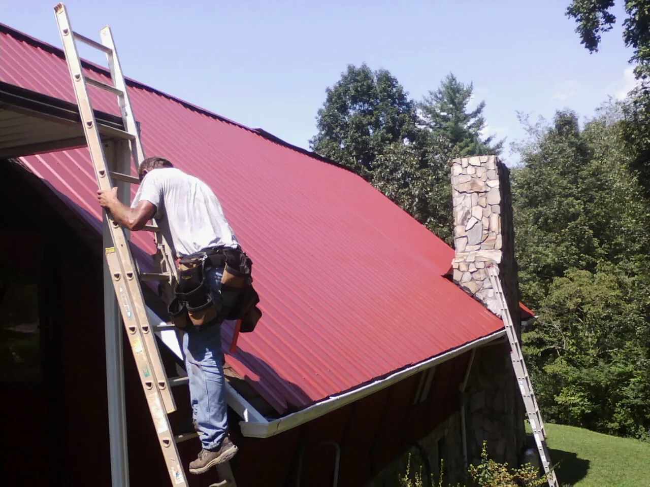 Roofer on a ladder beside a red metal roof and stone chimney — Soffit & Fascia Repair by Roofing Brothers in Mountainville